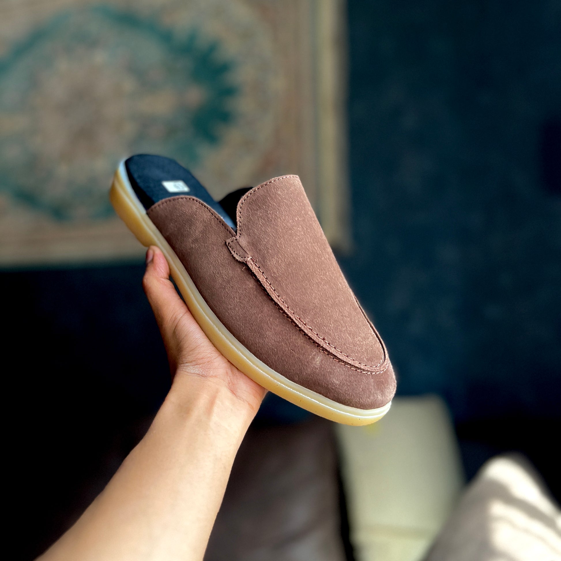 Brown suede slip-on shoe held by a hand with a blurred background
