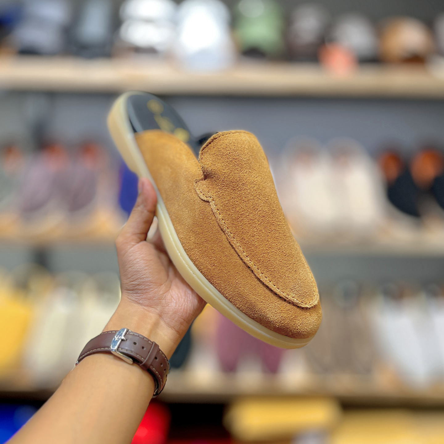 A tan suede slip-on loafer shoe held up by a hand in a shoe store, showing the side profile of the shoe with its cream-colored sole and dark interior.