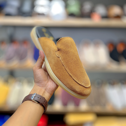 A tan suede slip-on loafer shoe held up by a hand in a shoe store, showing the side profile of the shoe with its cream-colored sole and dark interior.